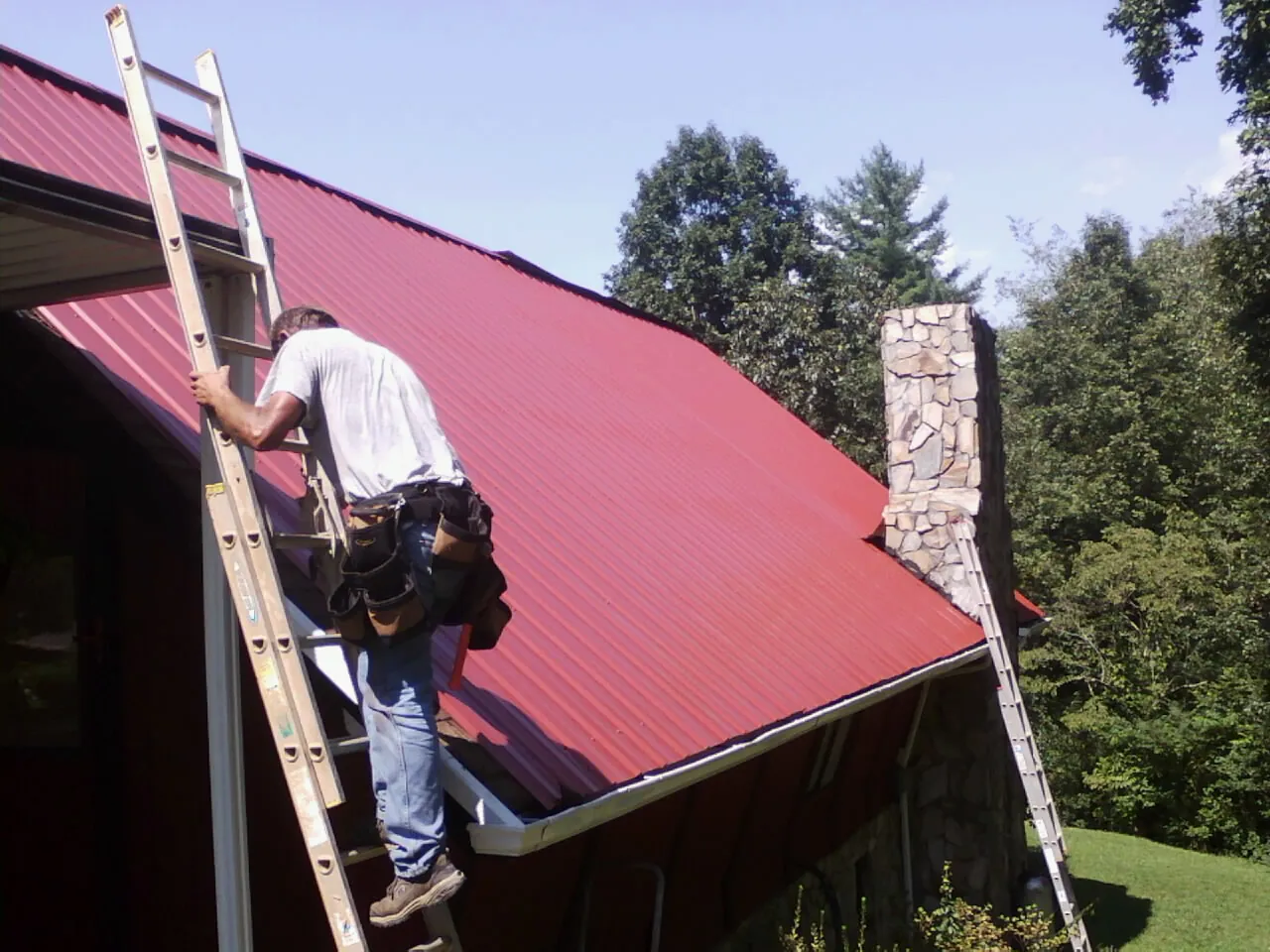 Roofer on a ladder beside a red metal roof and stone chimney — Soffit & Fascia Repair by Roofing Brothers in Quinlan